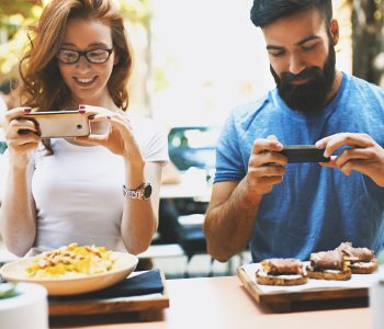 Couple taking shots of food. Closeup front view of a couple at a restaurant taking shots of their meal in order to post it online. It's seems very strange if you do not do this nowadays.